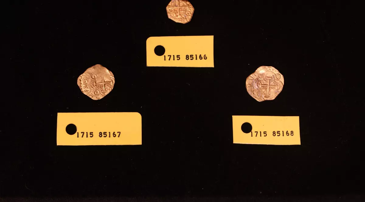 Imaginea prezintă trei monede vechi din aur sau argint, de formă neregulată și cu semne slab vizibile (posibil cob-uri spaniole), așezate pe un fundal negru. Fiecare monedă este însoțită de o etichetă galbenă pe care este scris un număr de inventar sau de identificare care începe cu 1715, sugerând o legătură cu faimoasa flotă spaniolă scufundată în acel an.