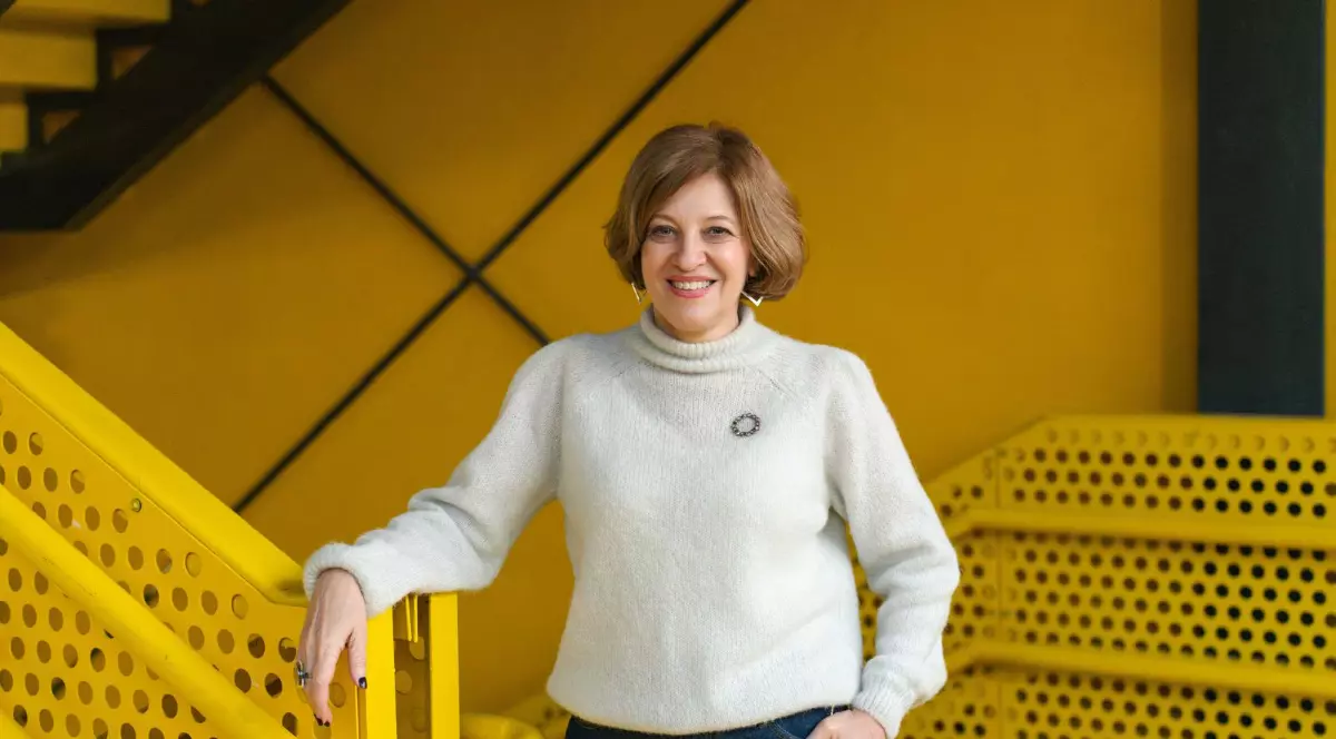 O femeie zâmbitoare, cu părul scurt și șaten, este surprinsă în fața unui fundal vibrant, dominat de culoarea galben intens și structuri metalice perforate. Este îmbrăcată cu un pulover gros, de culoare deschisă, cu guler pe gât și blugi, sprijinindu-se cu o mână pe balustrada galbenă a unei scări.