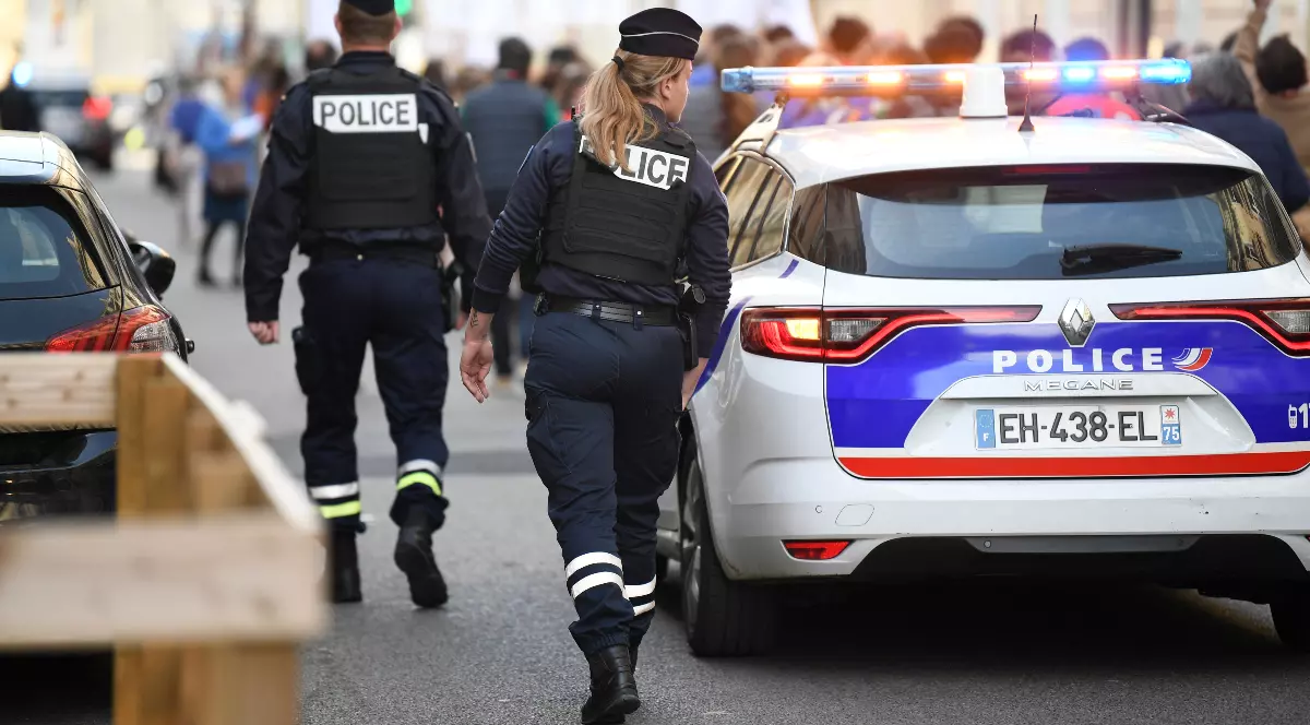 Cei doi polițiști francezi (un bărbat și o femeie), îmbrăcați în uniforme închise cu veste tactice inscripționate "POLICE", se îndepărtează de o mașină de patrulă albă cu girofarurile aprinse. Ofițerii patrulează pe o stradă urbană, cu o mulțime de oameni neclară în fundal, sugerând prezența lor în timpul unui eveniment public.