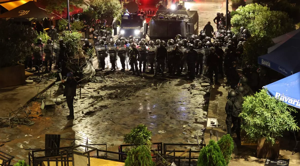 Imaginea surprinde o scenă de tensiune nocturnă, unde un rând compact de forțe de ordine (posibil poliție sau jandarmerie) în echipament de intervenție se confruntă cu o zonă dezordonată și plină de resturi. În prim-plan se văd mese și scaune răsturnate, iar pe fundal, două vehicule blindate și un grup mare de agenți menținând o linie de apărare, sugerând o intervenție după tulburări civile sau un protest.