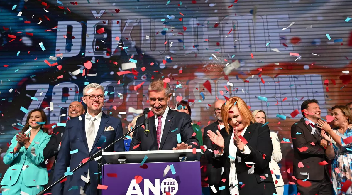 Imaginea surprinde un moment de sărbătoare politică, cu un grup de lideri și susținători în picioare pe o scenă, în timp ce confetti roșii, albe și albastre cad peste ei. Un bărbat în centru vorbește la un pupitru purtând sigla partidului 