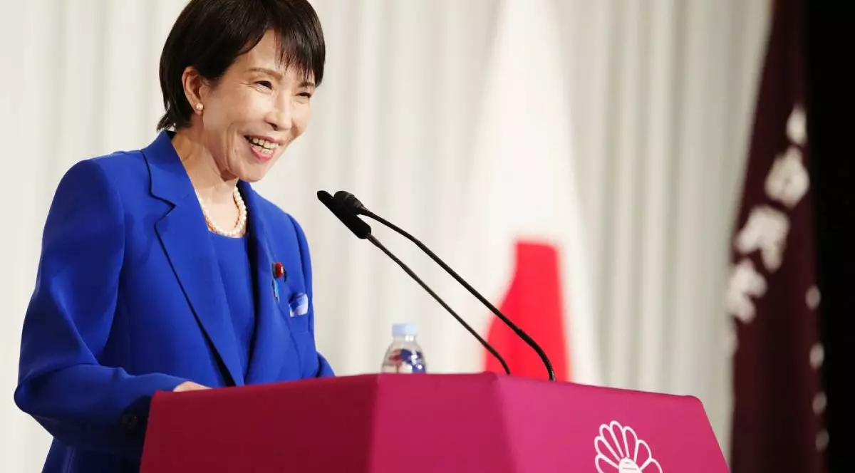 Sane Takaichi, prima femeie premier din istoria Japoniei, îmbrăcată într-un costum albastru, în timp ce vorbește la microfon la o conferință de presă, zâmbește și are buzele date cu ruj roșu