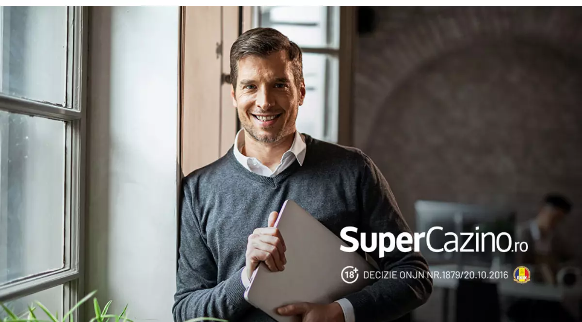 Barbat imbracat smart casual intr-o camasa alba si un pulover gri, subtire pe deasupra, tine in mana o tableta. Se afla intr-o camera care are peretele din spate de caramida, langa un geam