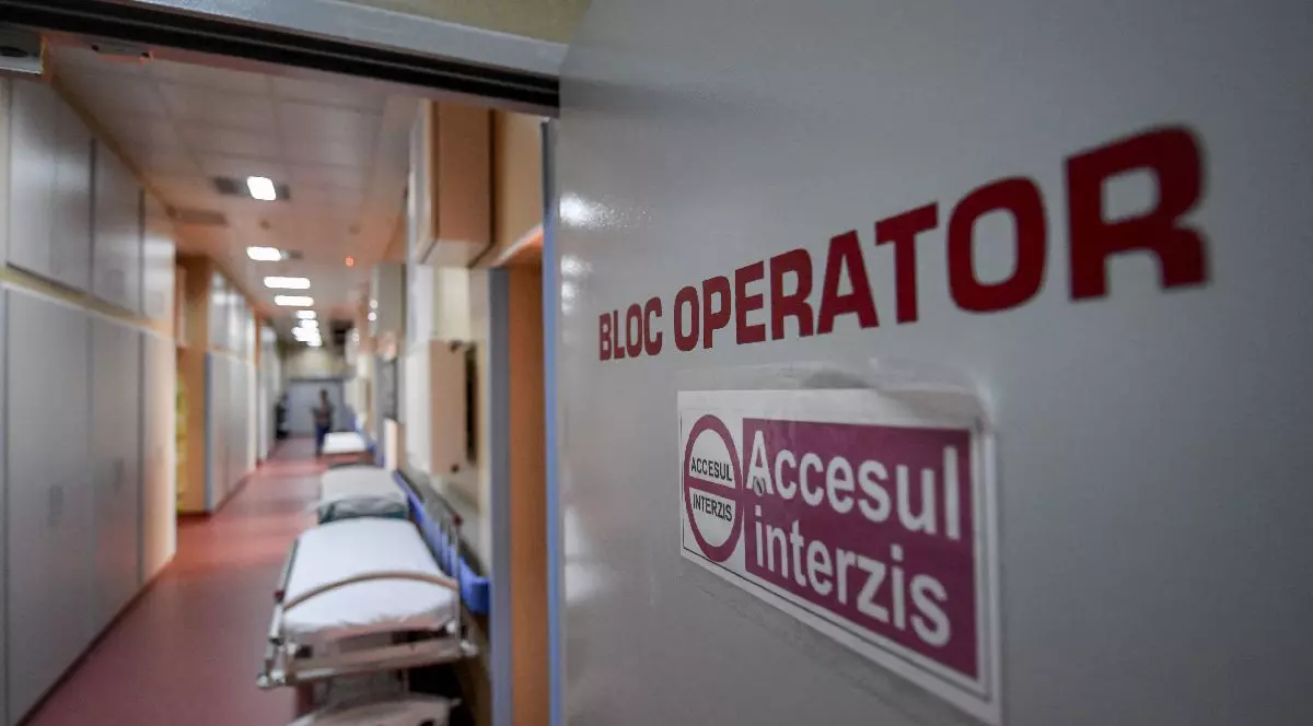 Paturi de spital sunt așezate pe un hol din spital, iar în prim-plan este o ușă dechisă pe care scrie bloc operator