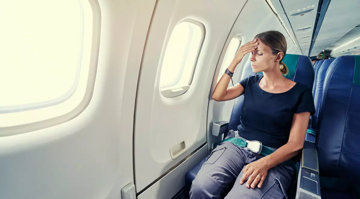 Femeie care sta in avion, cu centura pusa, isi tine mana la funte mimand o durere de cap