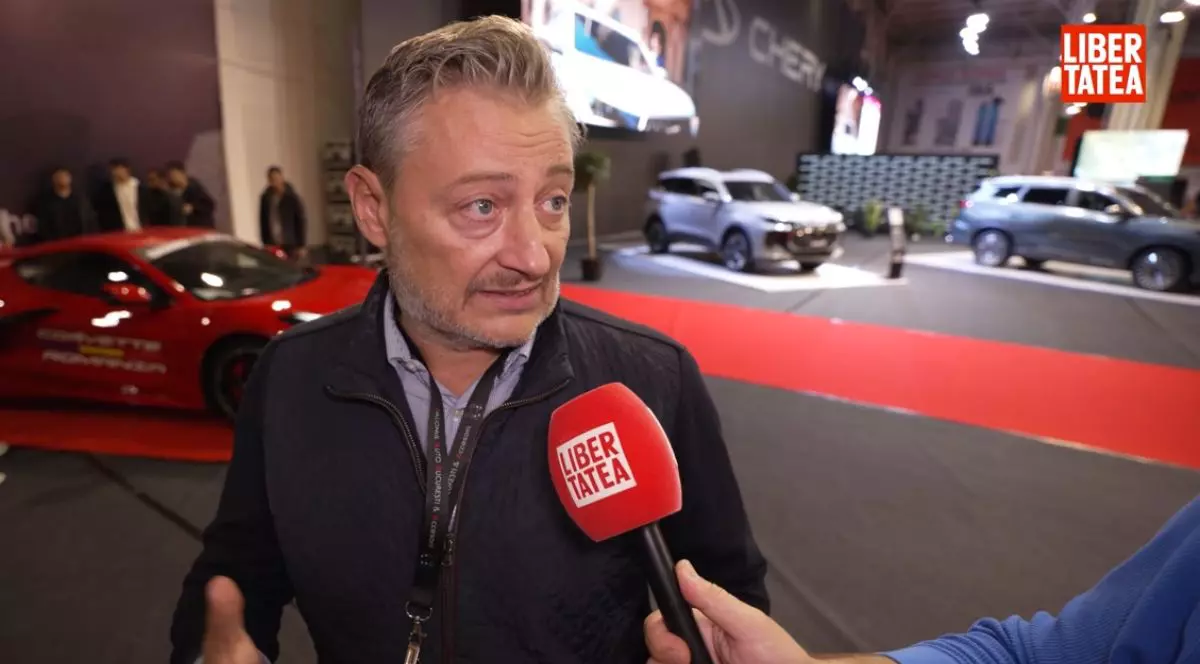 Sorin Rădăcineanu, organizatorul Salonului Auto București 2025, îmbrăcat cu un pulover negru peste o cămașă albastră, cu microfonul Libertatea în față, iar pe fundal standuri de mașini