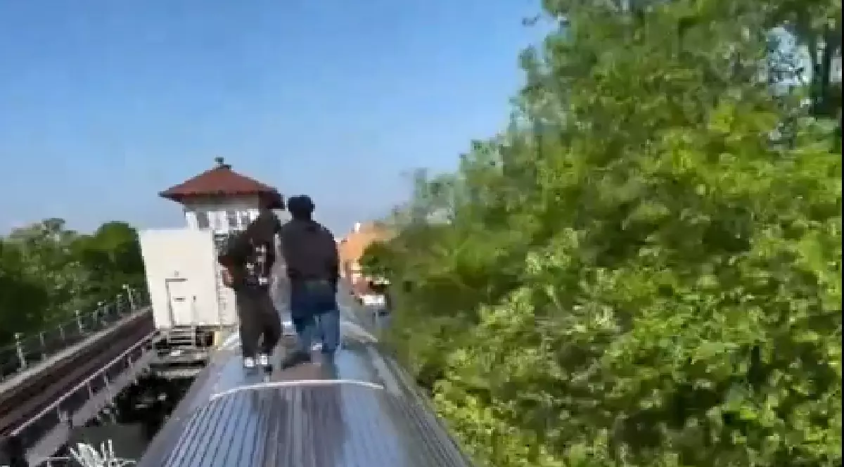 Imaginea surprinde un moment extrem de periculos în care două persoane stau în picioare pe acoperișul unui vagon de tren sau metrou în mișcare. Acțiunea, cunoscută sub numele de subway surfing (fără a promova acest comportament periculos), se desfășoară în aer liber, cu vegetație abundentă și o clădire de gară vizibile pe laterale.