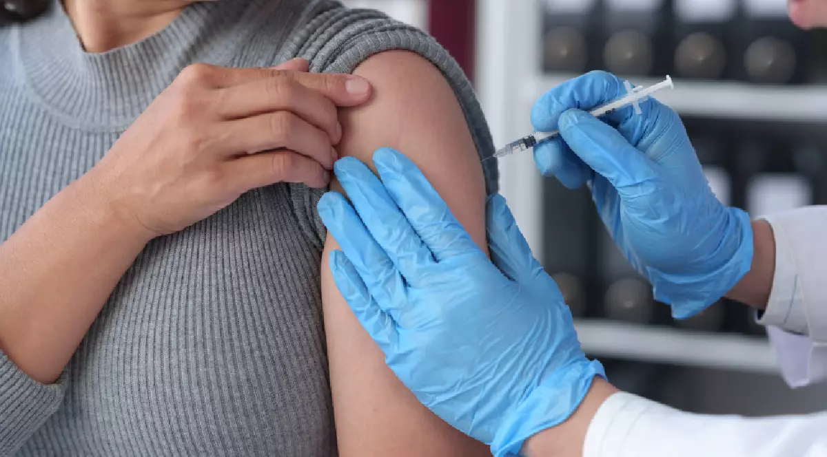 Imaginea surprinde de aproape momentul administrării unui vaccin sau a unei injecții într-un cadru medical. O persoană îmbrăcată în gri își ține brațul, în timp ce o mână a unui lucrător medical, protejată de mănuși albastre, injectează serul cu o seringă.