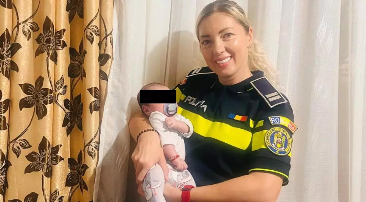 O polițistă blondă, îmbrăcată în uniformă, în timp ce ține un bebeluș în brațe și zâmbește în fața camerei.