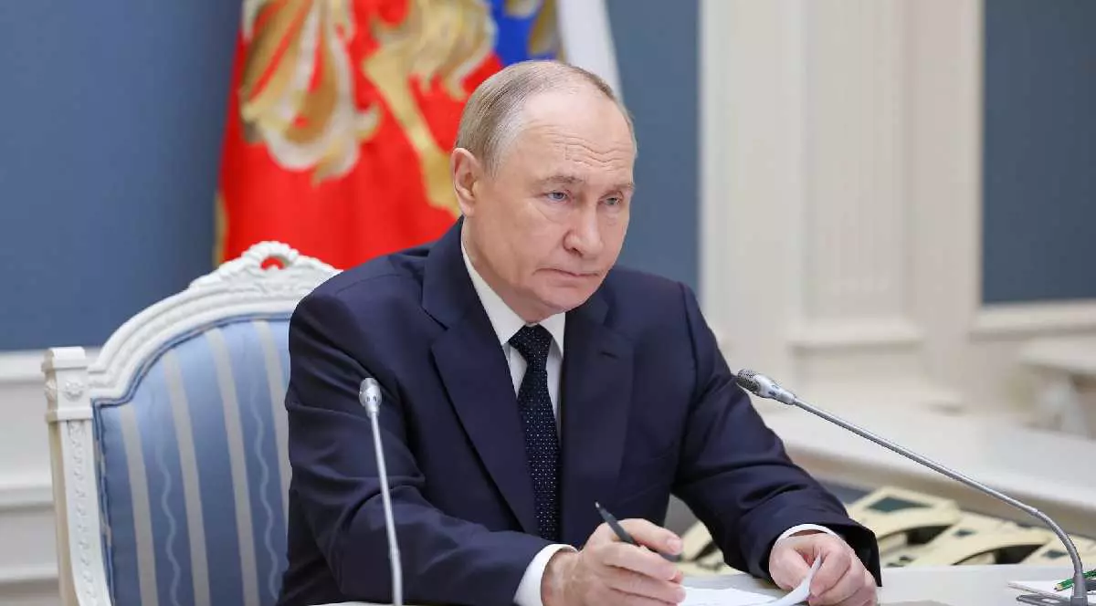 Vladimir Putin în biroul său, îmbrăcat într-un sacou negru, o cămașă albă și o cravată neagră, stând cu mâinile pe masă, ținând într-una din mâini un document iar în cealaltă un pix negru