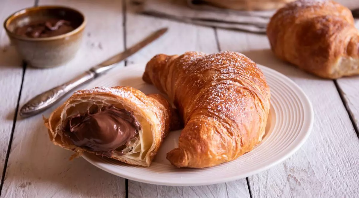 Croissante cu ciocolată pentru micul dejun sunt pe o farfurie albă care este pusă pe o masă rustică de lemn