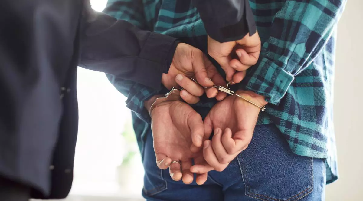 Cadru strans cu mainile unui polistist in timp ce incatuseaza un barbat cu o camasa in carouri verde cu negru si pantaloni de blugi