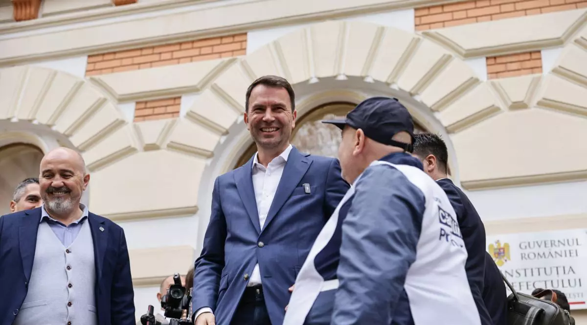 Cătălin Drulă îmbrăcat într-un costum bleumarin cu o cămașă albă pe scările Prefecturii București, înconjurat de susținători cu veste albe și șepci, la depunerea candidaturii pentru alegerile primarului general din București, 12 noiembrie 2025