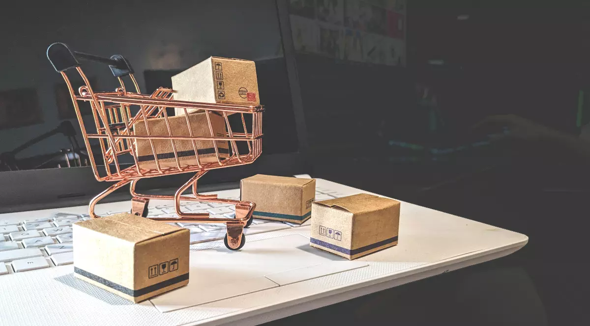 Un coș de cumpărături în miniatură pe un laptop, înconjurat de cutii mici de carton.