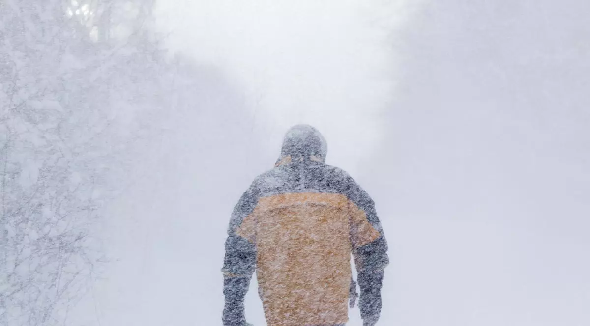 Un bărbat îmbrăcat într-o jachetă neagră cu galben, cu spatele la cameră, înconjurată de ninsoare abundentă, cu un fundal neclar, acoperit de zăpadă