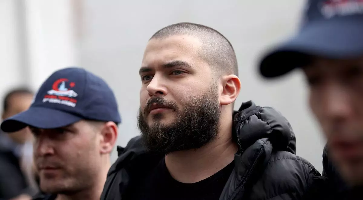 Imaginea prezintă un tânăr cu barbă și cap ras, îmbrăcat într-o jachetă neagră, înconjurat de doi bărbați cu șepci albastre