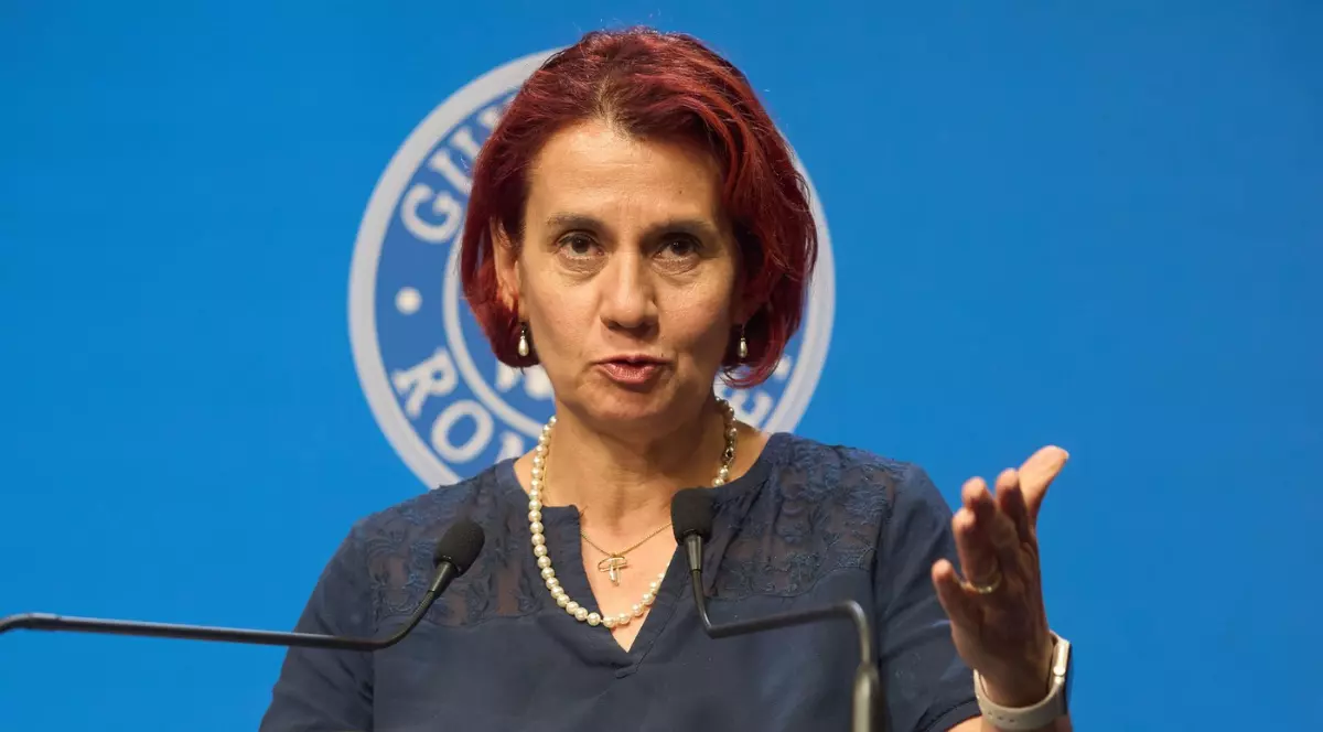 Imaginea prezintă un portret de jurnalistă, surprinsă în timp ce vorbește la un pupitru, având părul roșcat și purtând un colier de perle și o bluză de culoare închisă. Fundalul este un perete albastru pe care se distinge o siglă mare, parțial vizibilă, care pare să aparțină Guvernului României, sugerând o conferință de presă sau o declarație oficială.