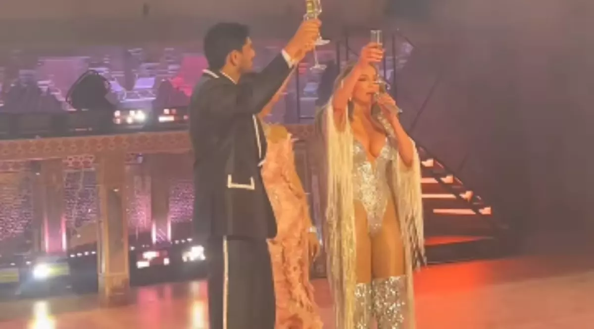 Imaginea o surprinde pe Jennifer Lopez pe scenă, îmbrăcată într-un body strălucitor, cu mâneci lungi și franjuri, ridicând un pahar de șampanie. Ea este însoțită de un bărbat în costum, care face același gest, iar fundalul pare a fi o scenă decorată extravagant.