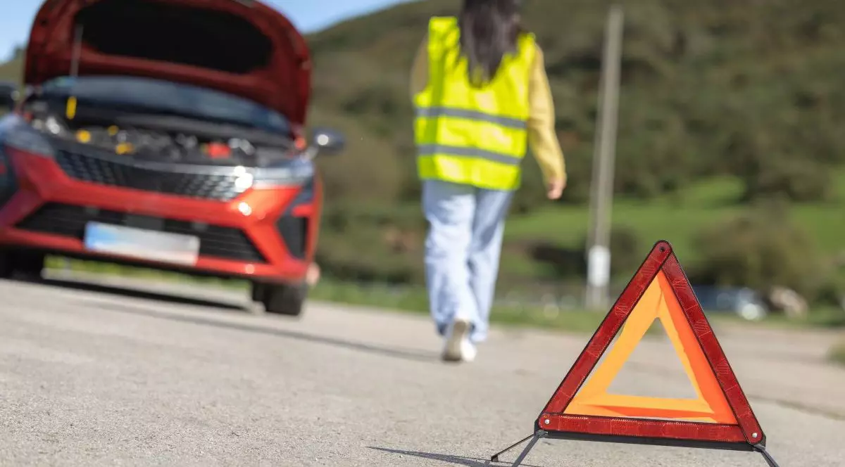 O tânără cu vestă reflectorizantă și fotografiată cu spatele merge spre o mașină toșie cu capota deschisă, iar în prim-plan este un triunghi roșu, reflectorizant, de urgență