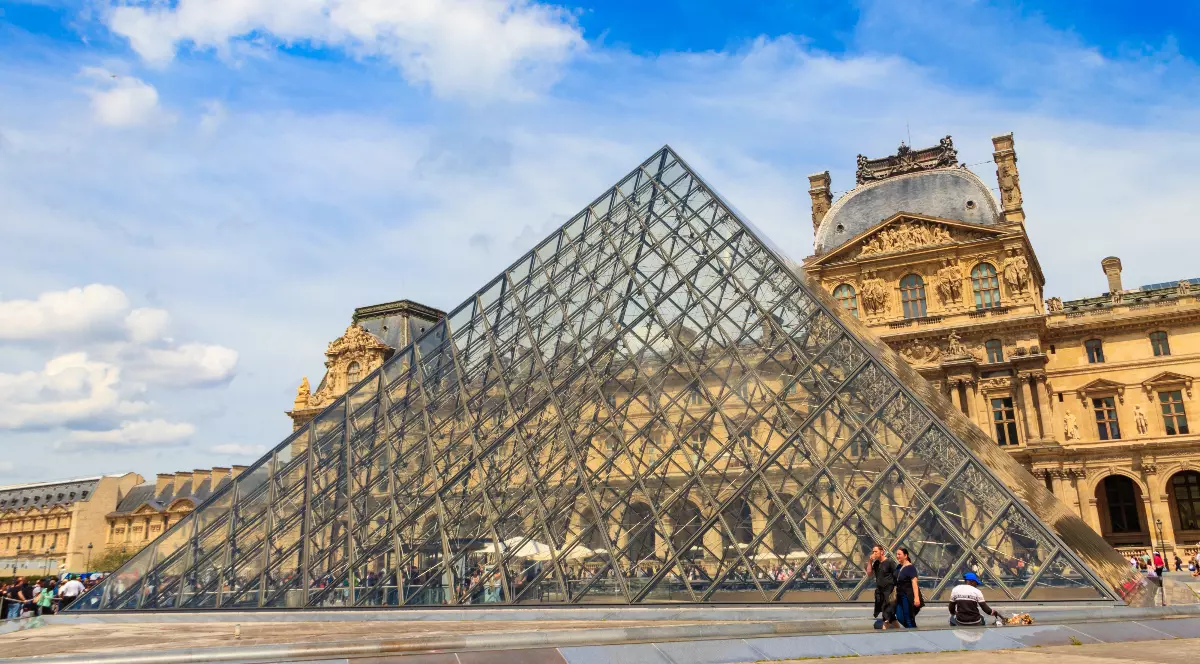 Imaginea prezintă celebra Piramidă de Sticlă din fața Muzeului Luvru din Paris, care contrastează puternic cu arhitectura clasică, masivă, a clădirilor din jur, sub un cer albastru cu nori puțini. Piramida modernă, cu structura sa metalică și panourile de sticlă, domină centrul cadrului, în timp ce la bază se pot observa câțiva vizitatori.