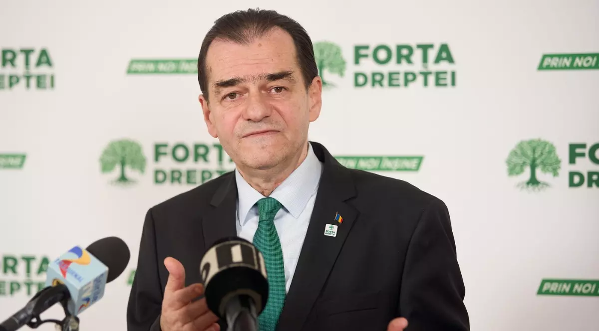 Ludovic Orban, îmbrăcat la costum, se adresează presei cu un gest din mâini. În spatele său se vede clar panoul cu logo-ul partidului Forța Dreptei, indicând o conferință de presă oficială.