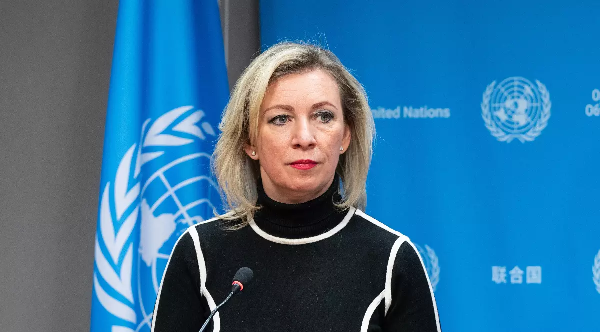 Maria Zaharova, purtând o bluză neagră cu guler înalt și detalii albe, în timp ce vorbește la un pupitru în cadrul unei conferințe. Fundalul este de culoare albastră intensă, fiind vizibil un steag al Națiunilor Unite și logo-ul în dreptul ei.