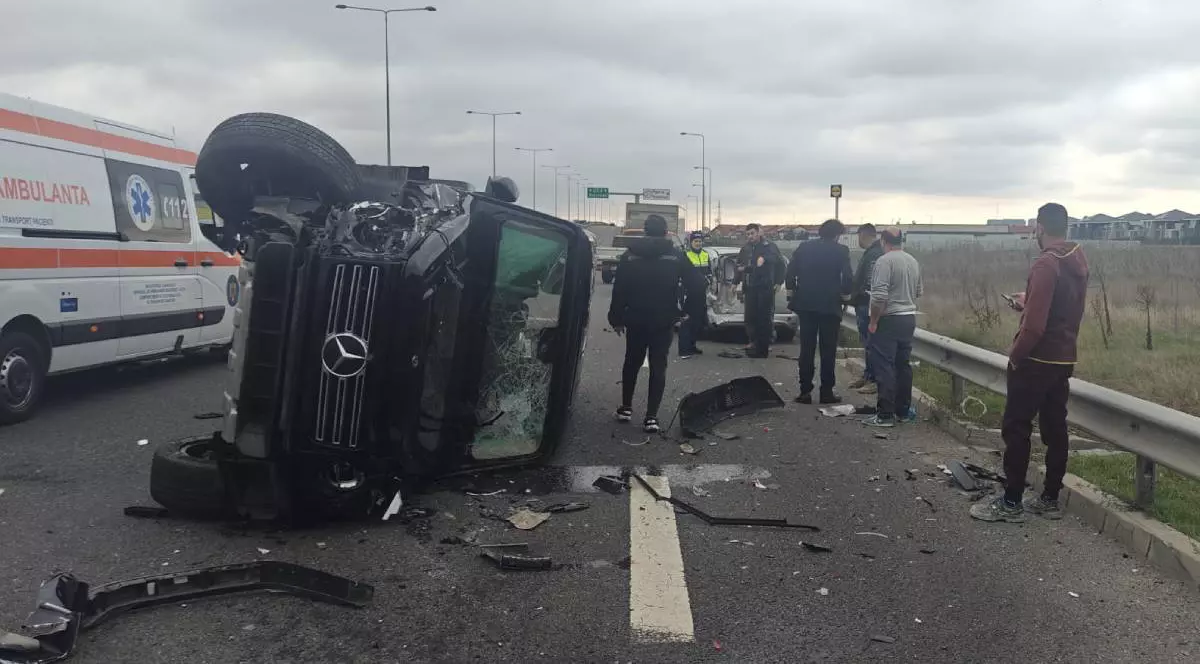 Un Mercedes G Clas negru, răsturnat pe șosea, are parbrizul spart și oamenii stau în jurul lui și se uită la mașină