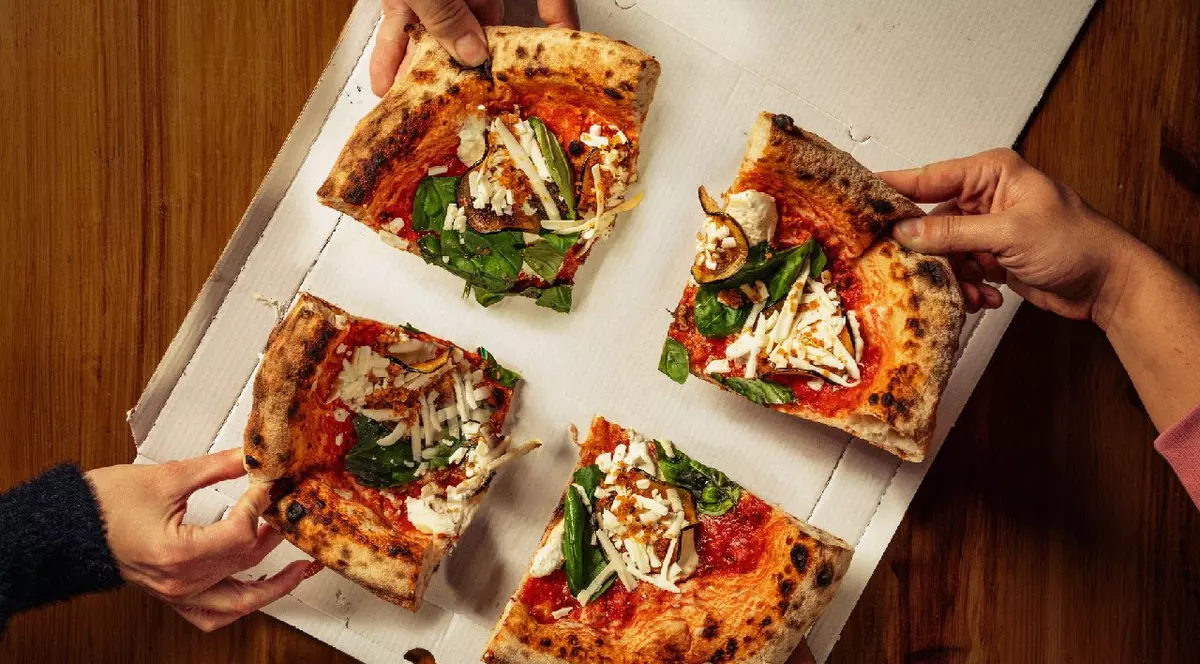 Patru mâini care iau felii de pizza cu brânză, busuioc și sos de roșii dintr-o cutie de pizza așezată pe o masă de lemn.