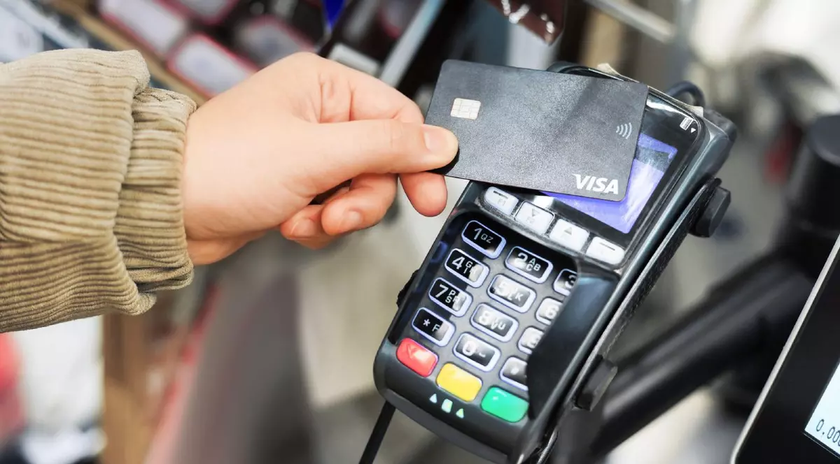 Mâna unui client ține un card de debit Visa negru lângă un terminal de plată pentru o tranzacție de plată fără contact.