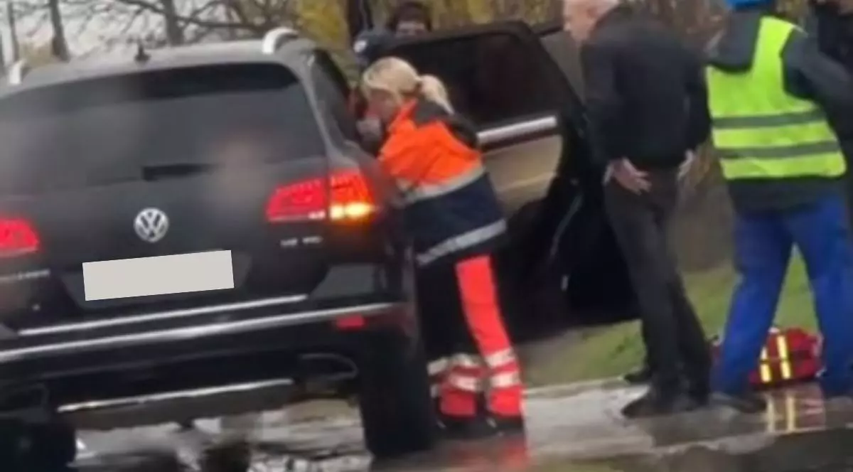 medic ambulanțier îmbrăcat într-o uniformă bleumarin cu portocaliu, stând aplecată spre interiorul unei mașini Volkswagen Touareg cu ușa deschisă, acordând primul ajutor