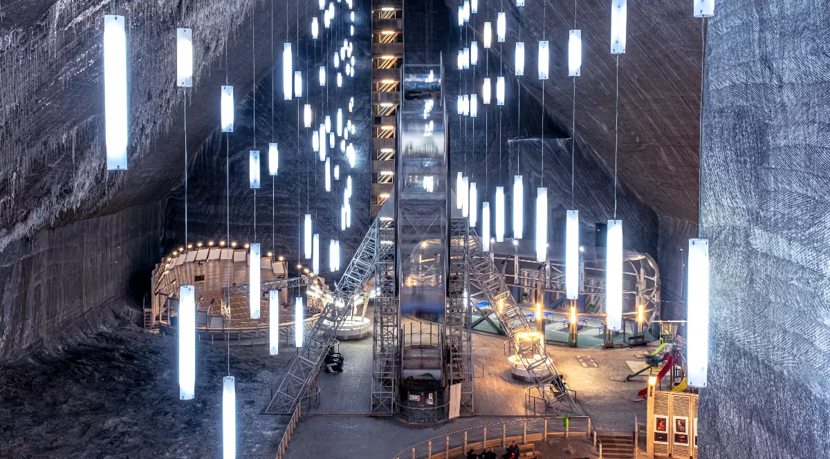 Imaginea suprinde interiorul spectaculos al Salinei Turda, cu pereții masivi din sare naturală și texturată. Spațiul larg, subteran, este iluminat modern de o multitudine de neoane verticale suspendate de tavan, care ghidează privirea către un lift central și zona de agrement de la baza camerei.