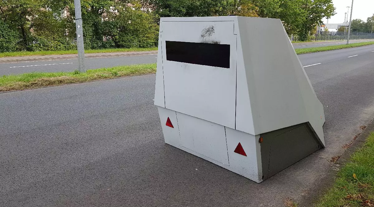 Imaginea prezintă un radar de viteză alb, dreptunghiular, amplasat pe o stradă