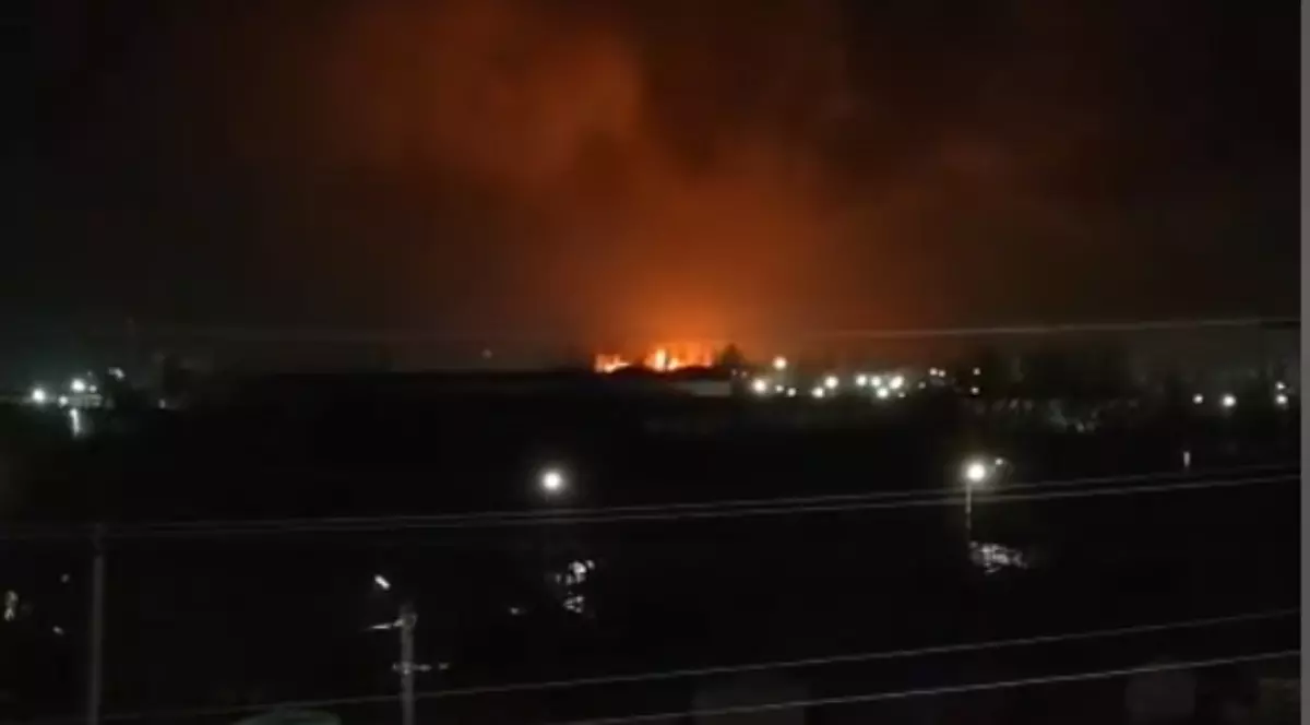 Imaginea surprinde o scenă nocturnă, dominată de un incendiu masiv vizibil la orizont, a cărui flacără roșiatică luminează intens cerul întunecat. În planul apropiat, se pot distinge siluetele unor clădiri și felinare stradale, care contrastează puternic cu lumina portocalie a focului din depărtare.