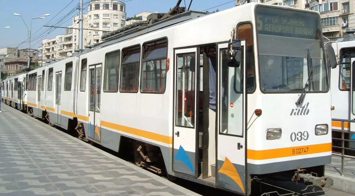 Imaginea prezintă un tramvai de culoare albă, cu bandă portocalie-galbenă, oprit într-o stație aglomerată din București. Pe afișajul frontal este indicat clar Traseul 5, cu destinația "P-ȚA SF. GHEORGHE / AEROPORT BĂNEASA", având în fundal clădiri de locuințe.