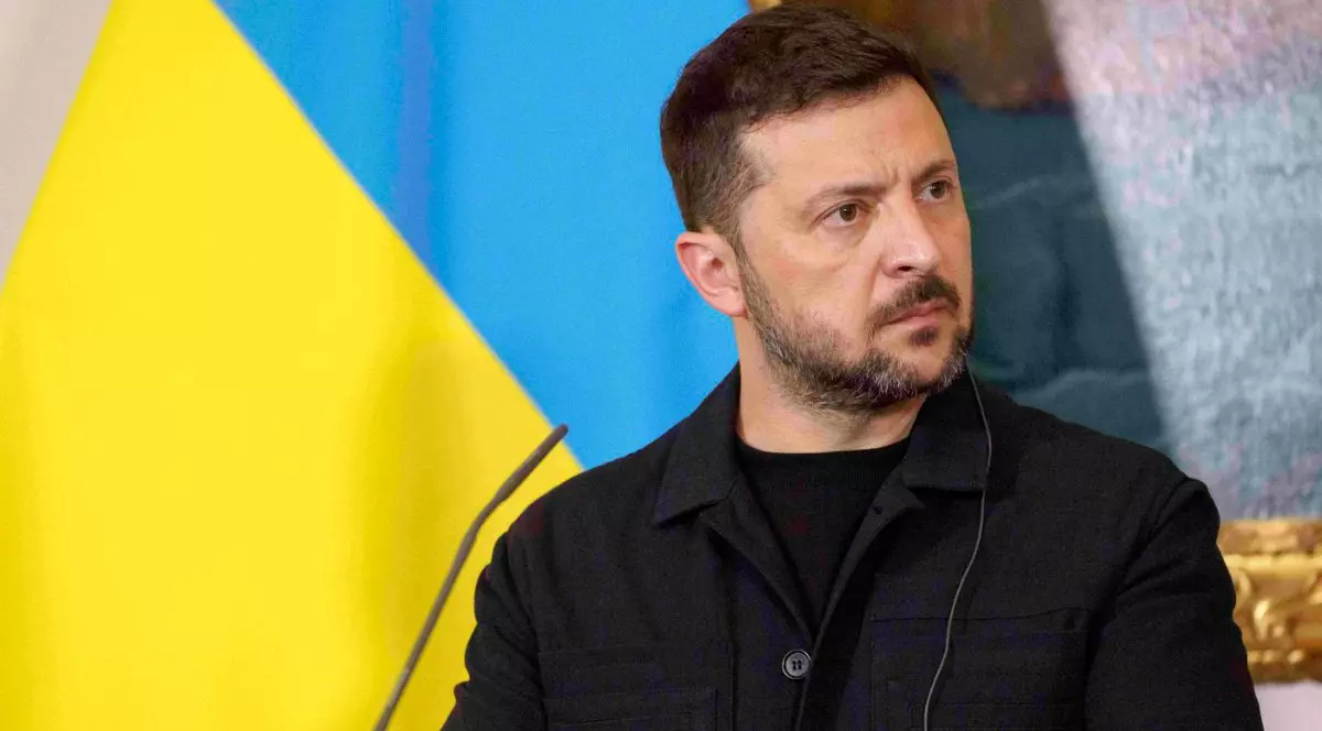 Imaginea este un prim-plan al președintelui ucrainean Volodimir Zelenski, care privește lateral cu o expresie serioasă, în timpul unei conferințe de presă sau discurs. El este îmbrăcat într-o ținută militară de culoare închisă, iar în spatele său se distinge clar steagul Ucrainei, în culorile galben și albastru.