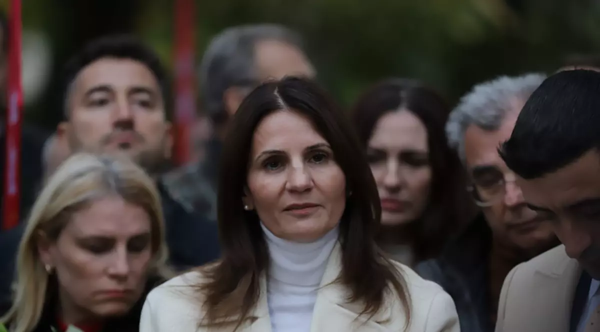 În imagine apare o femeie îmbrăcată într-un palton deschis la culoare, aflată în prim-plan, cu expresie serioasă și concentrată. Poartă un pulover alb pe gât și are un microfon prins pe haină, semn că este probabil în timpul unei declarații publice.