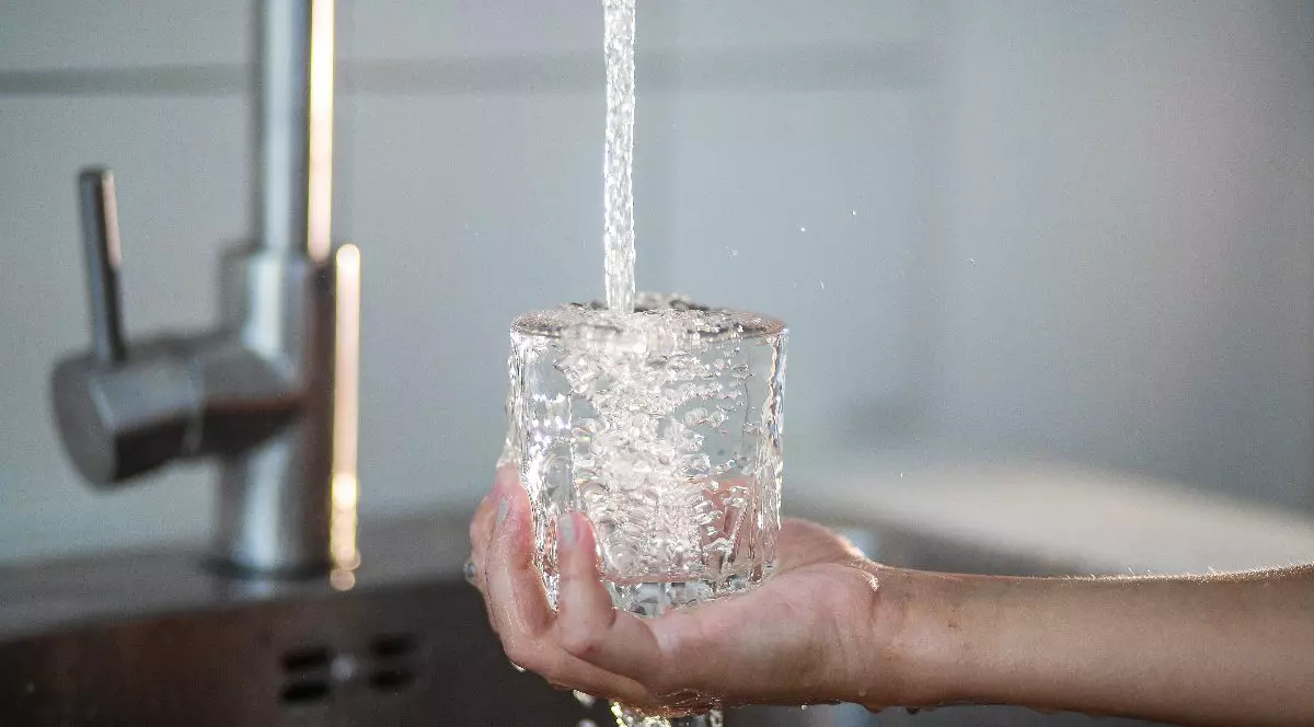 În imagine este surprins un pahar ținut sub jetul de apă de la chiuvetă, care se umple până dă pe dinafară. Fundalul este simplu, evidențiind gestul și claritatea apei care curge.