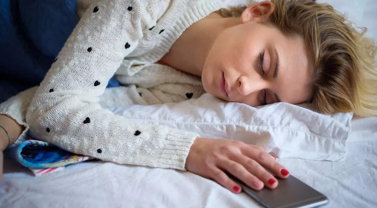 O tânără blondă, cu un pulover alb cu inimioare negre, doarme pe un pat pe care este un cearșaf alb și ține o mână pe telefonul mobil