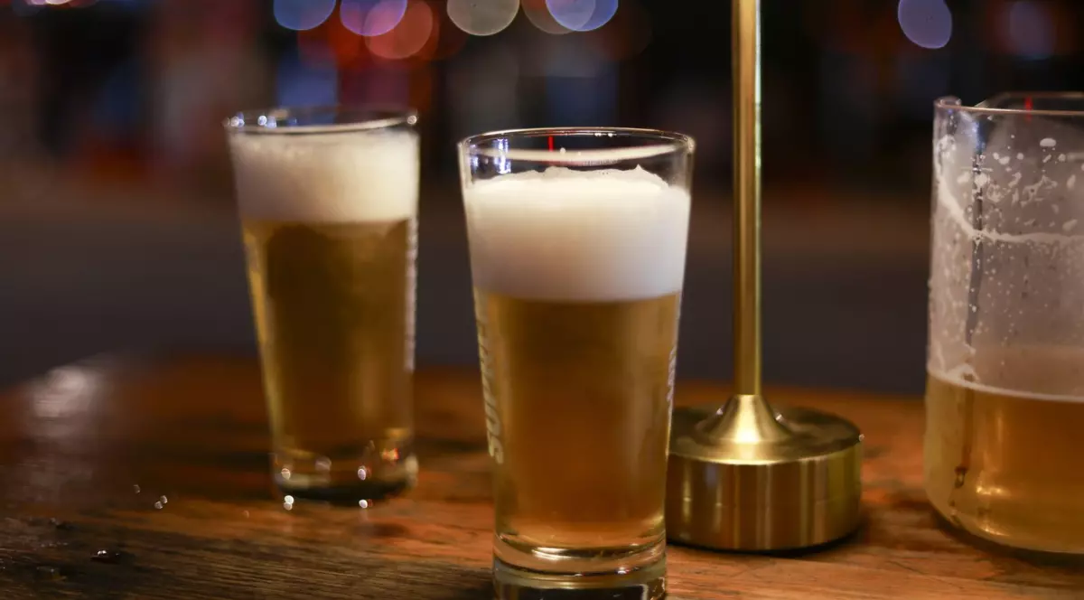 În imagine se văd mai multe pahare cu bere blondă, proaspăt turnată, cu spumă densă deasupra, așezate pe o masă din lemn. Fundalul este estompat, cu lumini difuze, sugerând atmosfera unui bar sau a unei terase pe timp de seară.