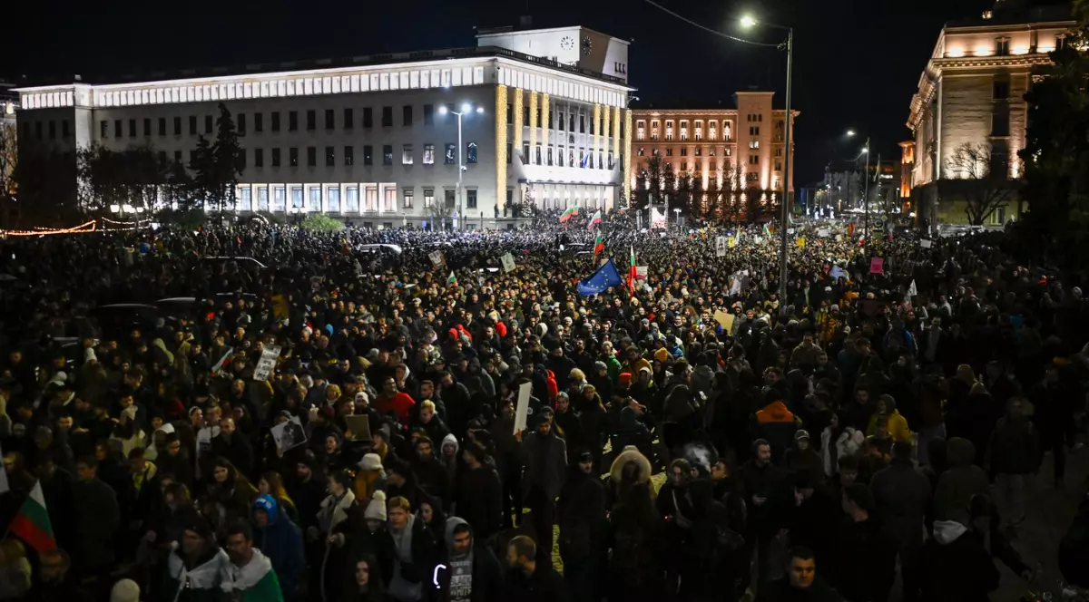 Imaginea arată o mulțime uriașă de protestatari adunați noaptea într-o piață din centrul unui oraș, fluturând steaguri și pancarte. Clădirile instituțiilor publice din fundal sunt iluminate, evidențiind amploarea și intensitatea manifestației.