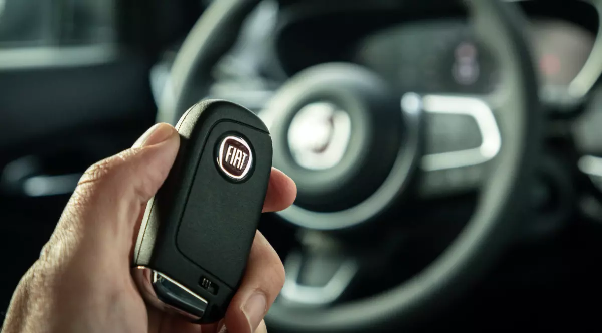 O persoană care stă în mașină și ține o cheie în mână pe care scrie FIAT. În fundal se vede un volan.