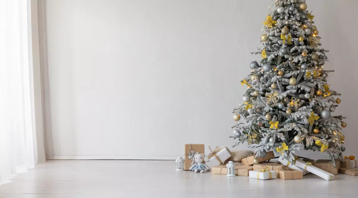 Imaginea surprinde un brad de Crăciun frumos împodobit, decorat cu globuri aurii și argintii, luminițe calde și fundițe. Sub brad se află cadouri împachetate elegant, asezate pe podea într-un decor minimalist, luminos și modern.