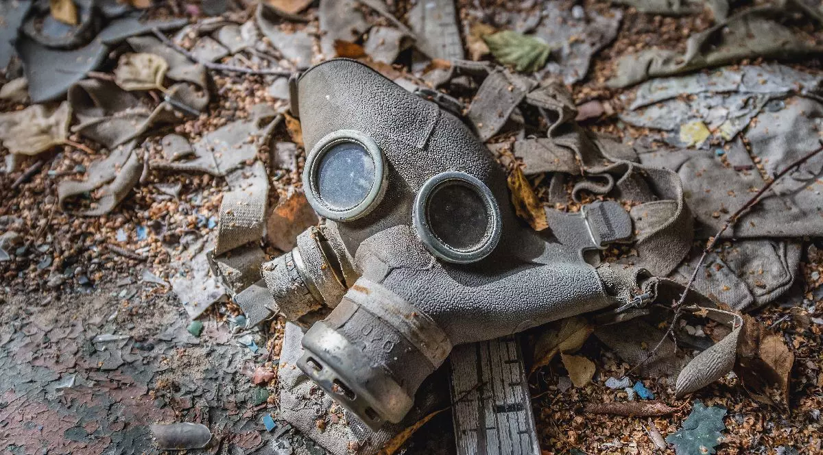 Prim-plan a unei măști de gaz vechi și uzate, zăcând pe un sol acoperit cu frunze uscate, crenguțe și pământ, sugerând abandonul sau degradarea.