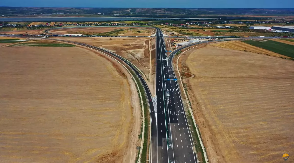Imaginea surprinde o secțiune din Drumul Expres Pitești–Craiova, văzută din dronă, cu sensuri de circulație bine delimitate și ramificații laterale. Înconjurat de câmpuri agricole, traseul rutier se întinde drept, evidențiind infrastructura modernizată în mijlocul unei zone rurale.