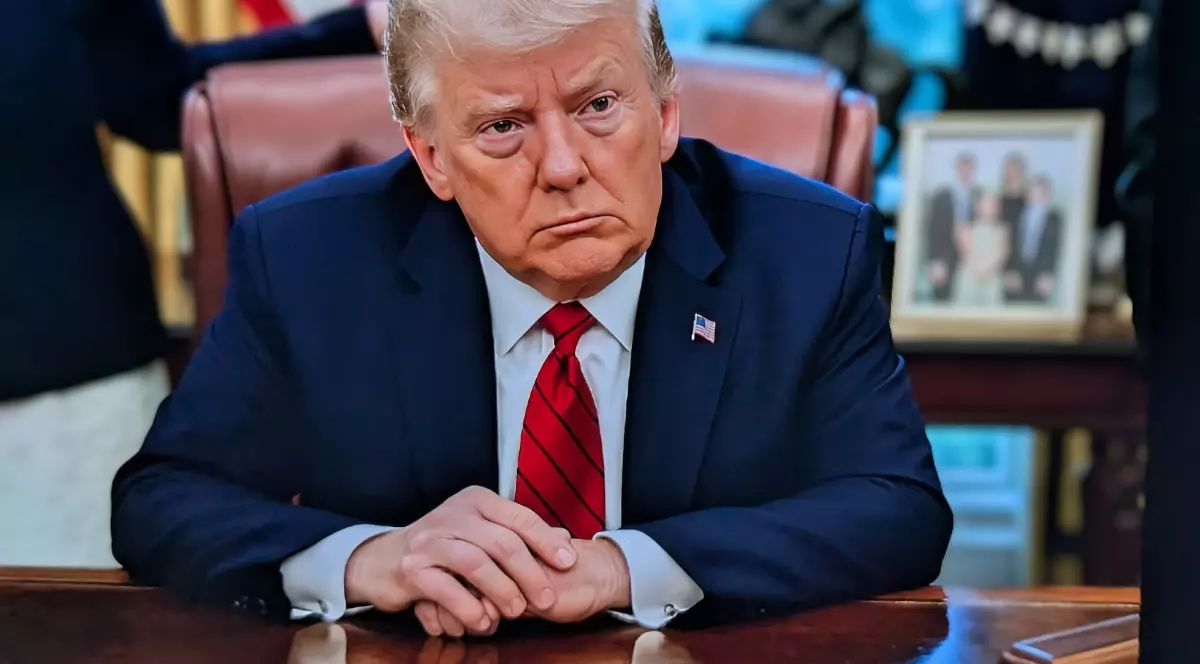 În imagine se vede un bărbat într-un costum închis la culoare, cu o cravată roșie, așezat la un birou. Are mâinile împreunate pe masă și o expresie serioasă pe chip. În spate se observă decor de birou oficial, inclusiv un steag și o fotografie de familie înrămată. Decorul sugerează un cadru formal, specific unui spațiu de conducere sau birou administrativ.