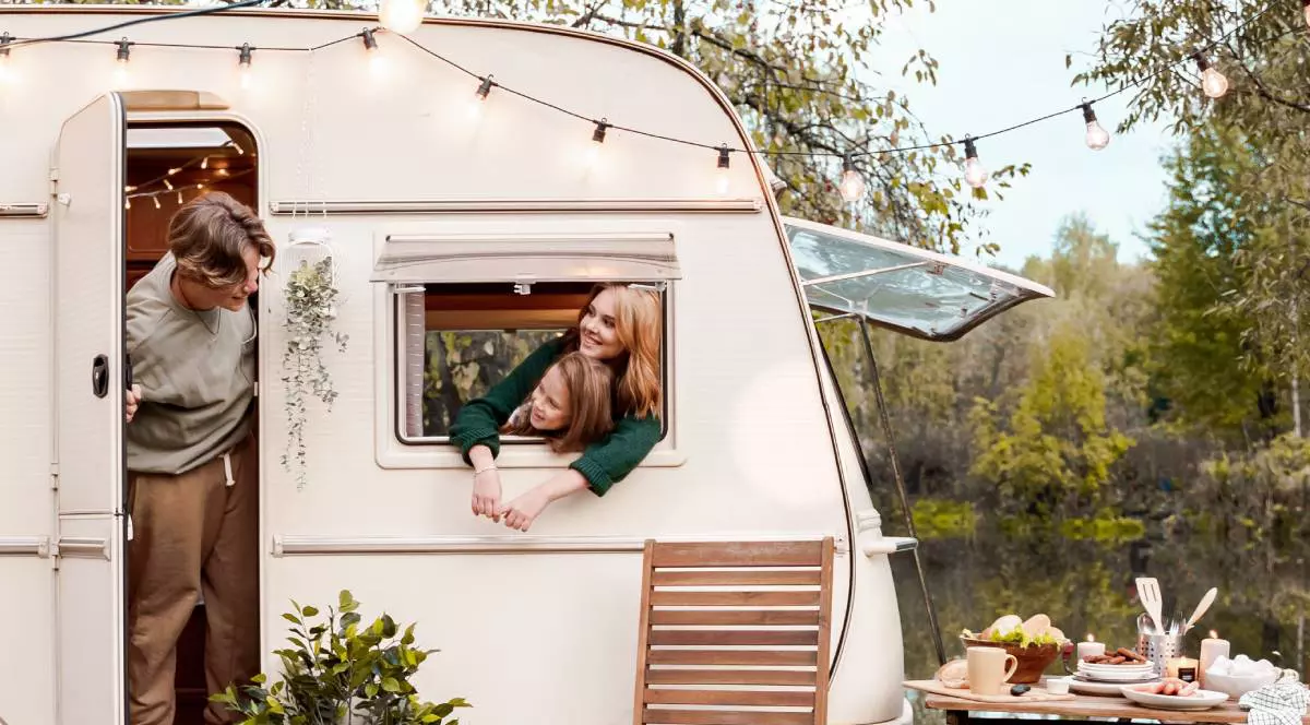 Imagine reprezentativa cu o familie cu o rulota frumos impodobita cu luminite. mama si o fetita au scos capul pe geam si se uita zambind la tatal care sta in usa deschisa a rulotei, in jurul lor copaci verzi si o masa frumos aranjata