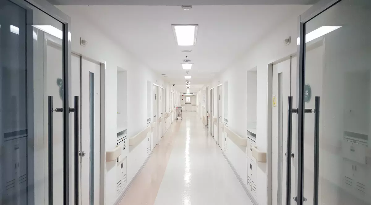 Imaginea surprinde un hol lung și luminos dintr-un spital, cu uși aliniate de-o parte și de alta și iluminare artificială uniformă. Spațiul este curat și ordonat, sugerând un mediu medical modern și lipsit de prezență umană în acel moment.