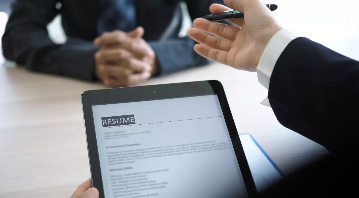 În imagine are loc un interviu de angajare, unde CV-ul unui candidat este analizat pe tabletă de către recrutor. În fața lui stă candidatul, cu mâinile împreunate, în timp ce intervievatorul îi adresează întrebări.