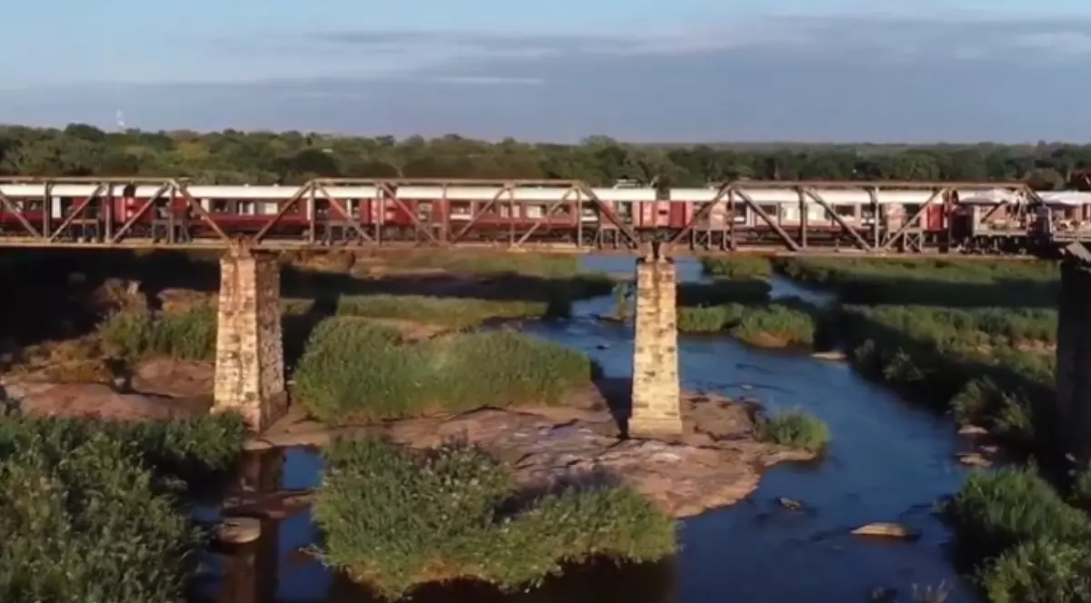 Imagine panoramică cu un șir de vagoane roșii cu alb, cu ferestre mari, alcătuind hotelul Kruger Shalati, staționat pe un pod metalic.