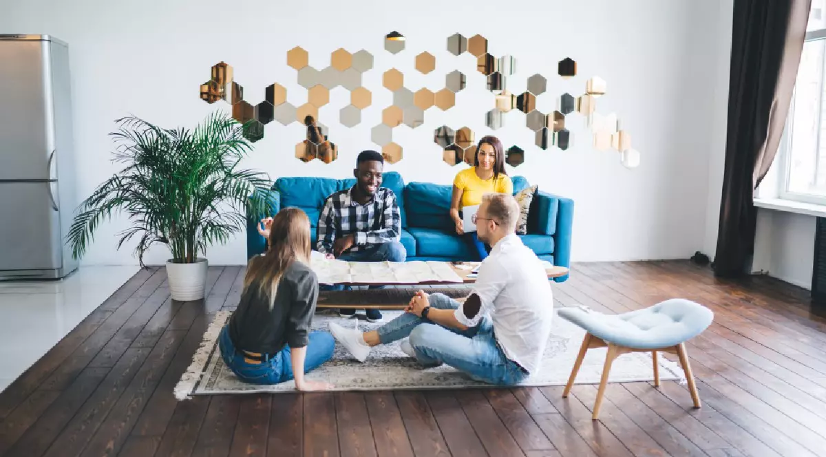 prieteni stând în jurul unei măsuțe de cafea acoperită cu o hartă și discutând idei despre călătorii împreună, în timp ce petrec timpul într-un studio confortabil, decorat cu plante și oglinzi pe pereți.